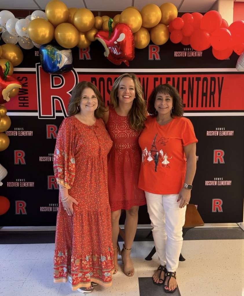 Photo of three women wearing red, smiliing, and standing in front of a black background with balloons.